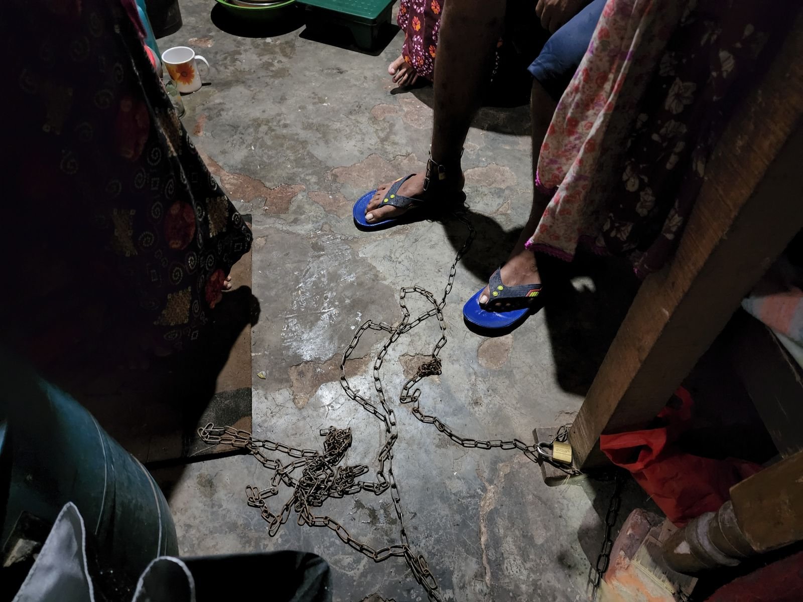 A heavy rusted metal chain coiled on a stained concrete floor with a padlock attached to a wooden post, surrounded by the feet and lower bodies of several people standing in a dark room