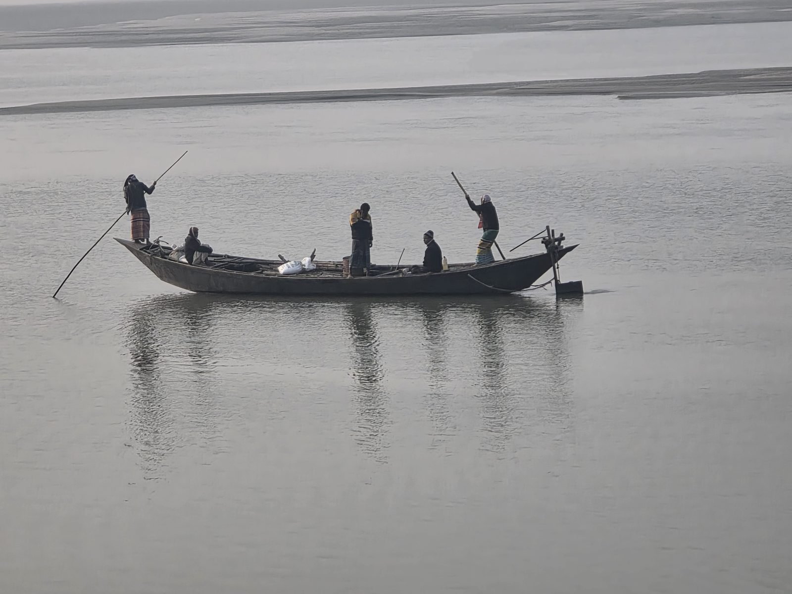 Five people on a long wooden country boat crossing a wide grey misty river at dawn — two boatmen standing at the prow and stern with long poles, three passengers seated in between, with a flat sandy riverbank visible in the pale background