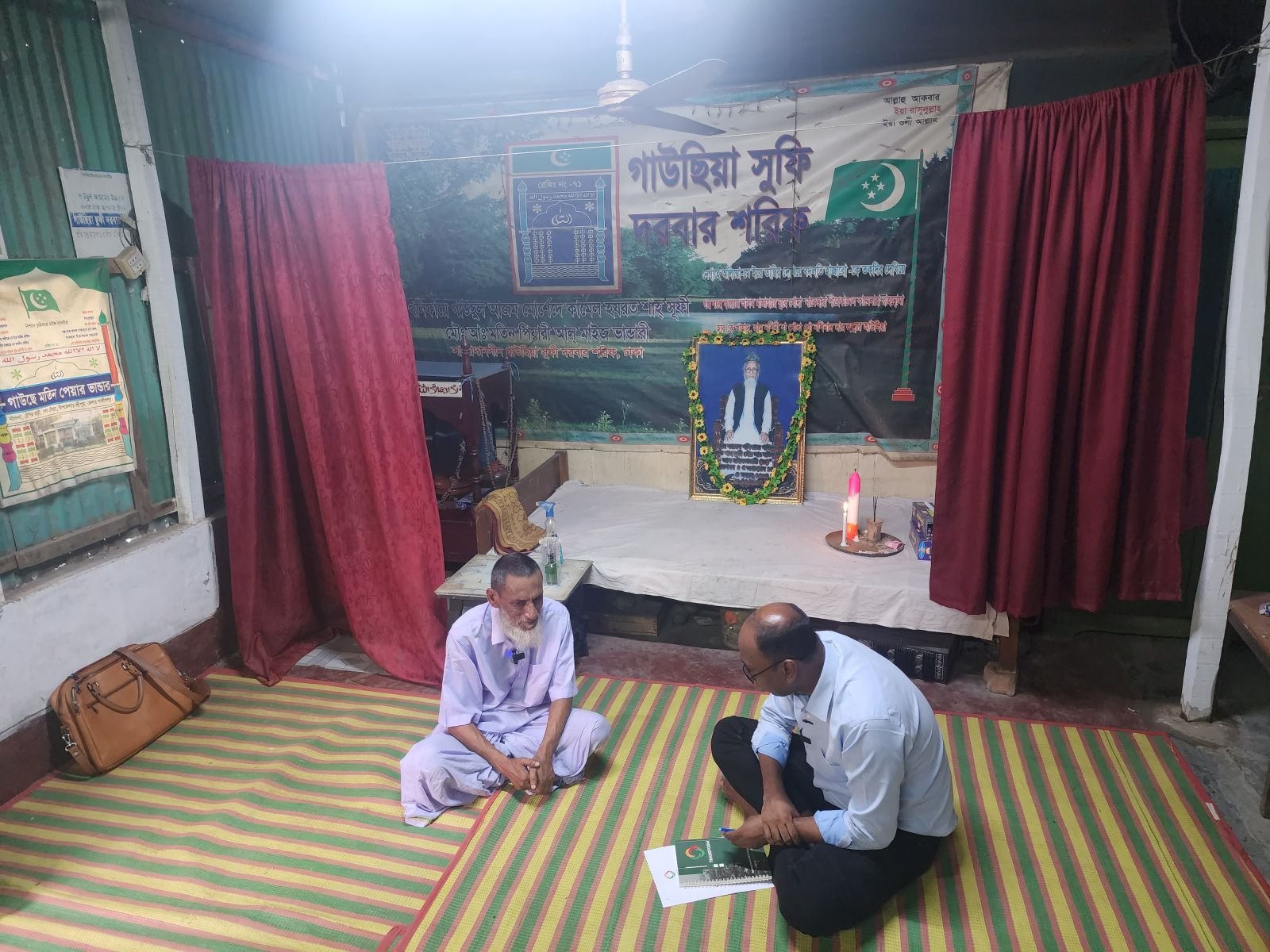 Two men sitting cross-legged on a striped woven mat inside a Sufi shrine — a younger man with a notebook facing an elderly man in white clothing with a white beard, with dark red curtains, a framed portrait, and a burning candle visible behind them on a low stage