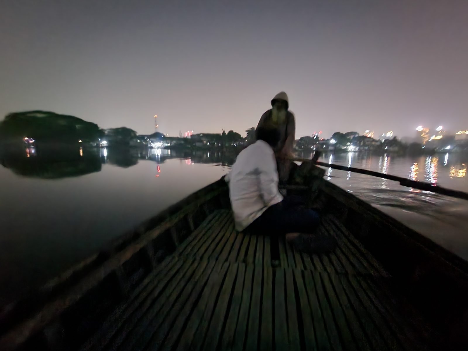 View from inside a bamboo raft at night, looking toward the bow where two silhouettes are visible — one seated and one standing with an oar — with soft city lights and their reflections visible across a dark river on both sides