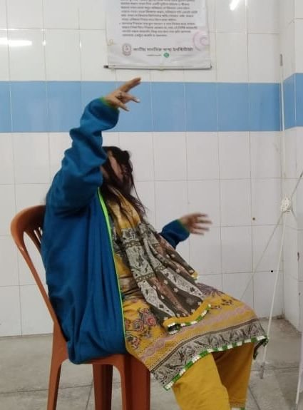 A woman in a bright blue jacket and yellow-printed clothes seated on an orange plastic chair, arms raised in opposite directions expressively — one pointing upward, one extending to the side — in a tiled institutional room with a Bengali poster on the wall