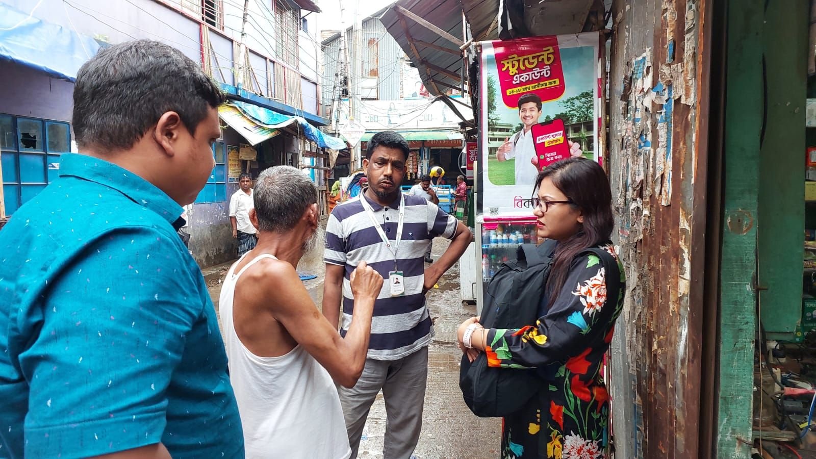 Three researchers — a man in a blue patterned shirt, a man in a striped polo with a lanyard, and a woman in a black floral outfit — speaking with an older man in a white vest in a narrow Dhaka alley lined with market stalls, a pink bank advertisement poster visible on a shop front