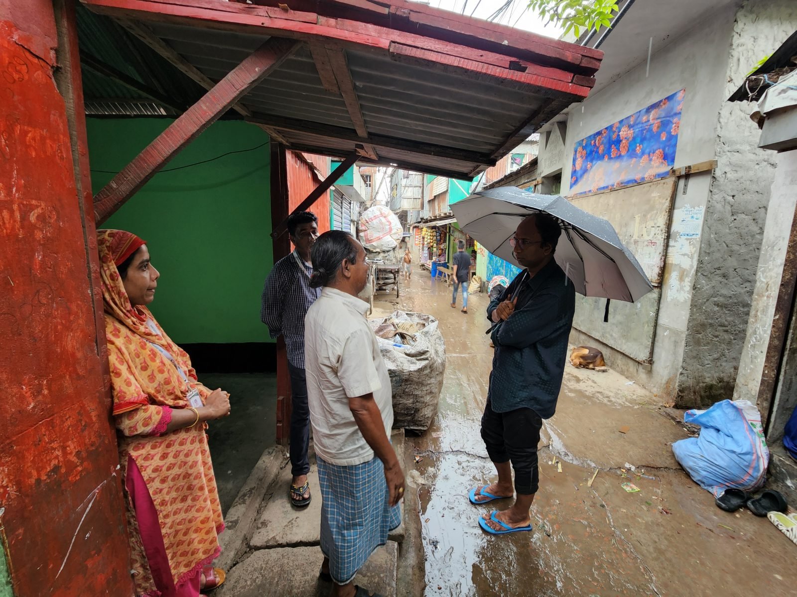 Four people sheltering from rain in a flooded Dhaka alley — a woman researcher in an orange floral dress with a lanyard stands against a red wall, a community member in a white lungi faces a male researcher in a dark shirt holding an umbrella, another researcher visible in the background