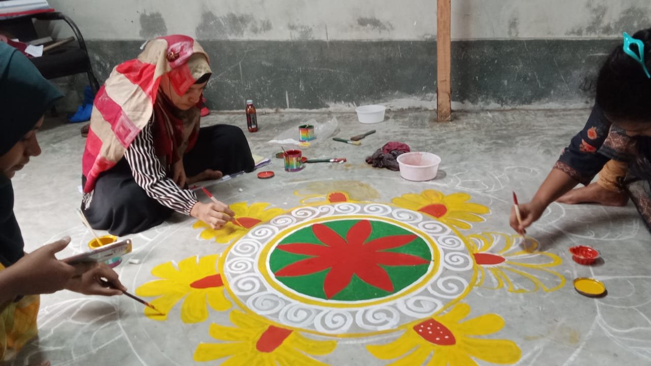 Two women seen from above working on a large floor mandala painting in red, yellow, green and white — a central circular flower motif surrounded by petal and scroll designs — with paint pots, brushes and a water bowl visible beside them