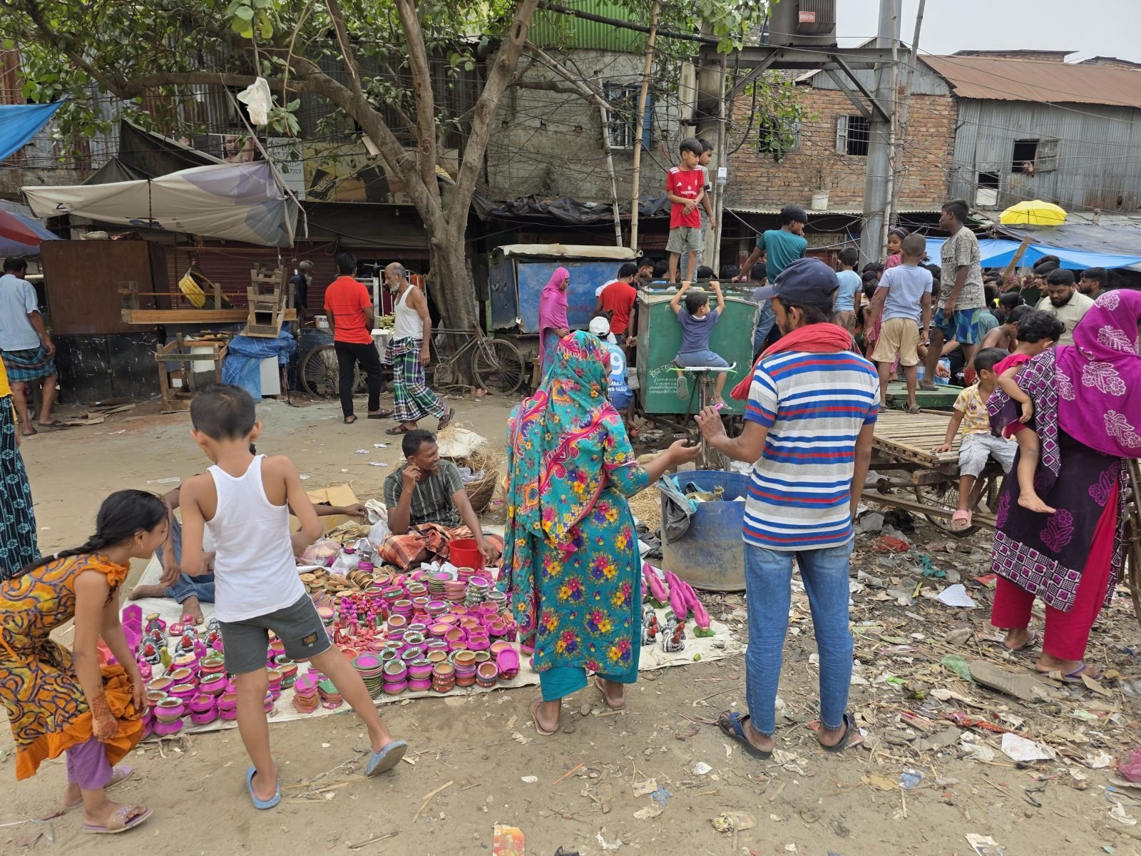 An outdoor market scene in Bangladesh with colourful pink, orange and red ceramics and baskets spread on the ground, a woman in a bright teal and floral sari examining goods, children nearby, and a crowd of shoppers behind them near ramshackle market stalls