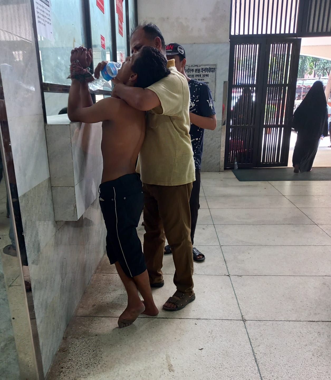 A shirtless barefoot young man with his hands bound behind his back in dark cloth is being embraced and supported by an older man in a khaki shirt at the tiled entrance lobby of a hospital, with a Bengali sign on the wall and a gated entrance behind them