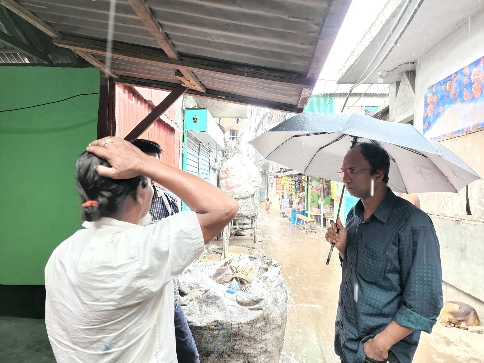 Two people standing in a narrow rain-soaked urban alley in Bangladesh — one sheltering from the heavy rain with a hand over their head, the other holding a large grey umbrella, facing each other in conversation, with colourful market stalls blurred behind them