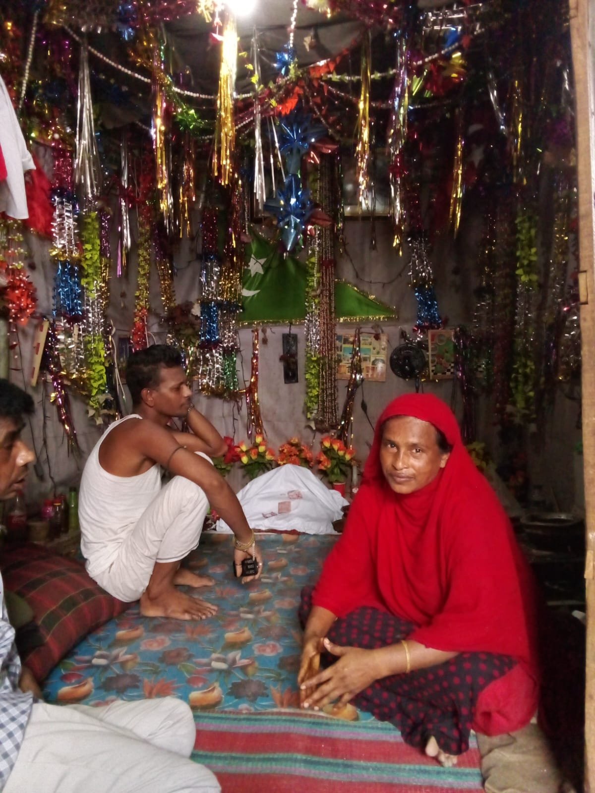Several adults seated on patterned floor mats inside a small room decorated with colourful metallic garlands and cloth
