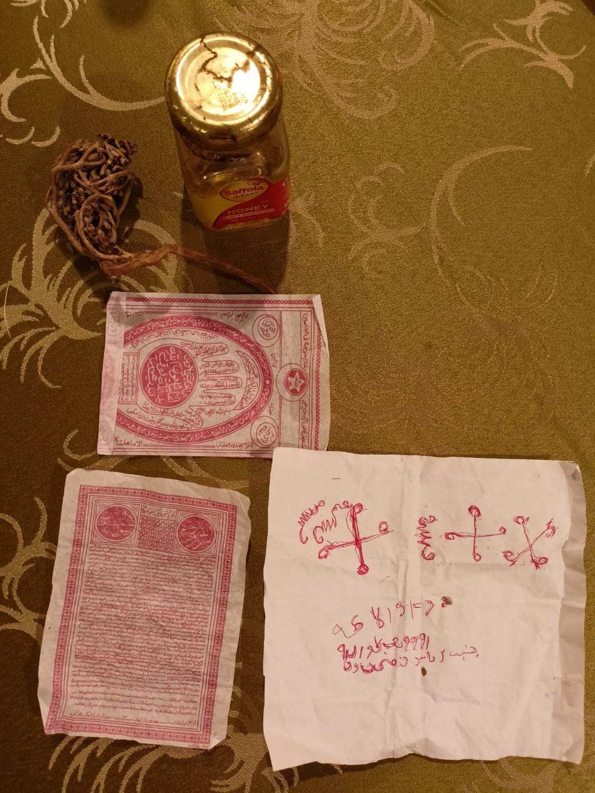 A glass jar, a knotted brown cord, two red printed papers and a white cloth marked with red symbols arranged on patterned fabric