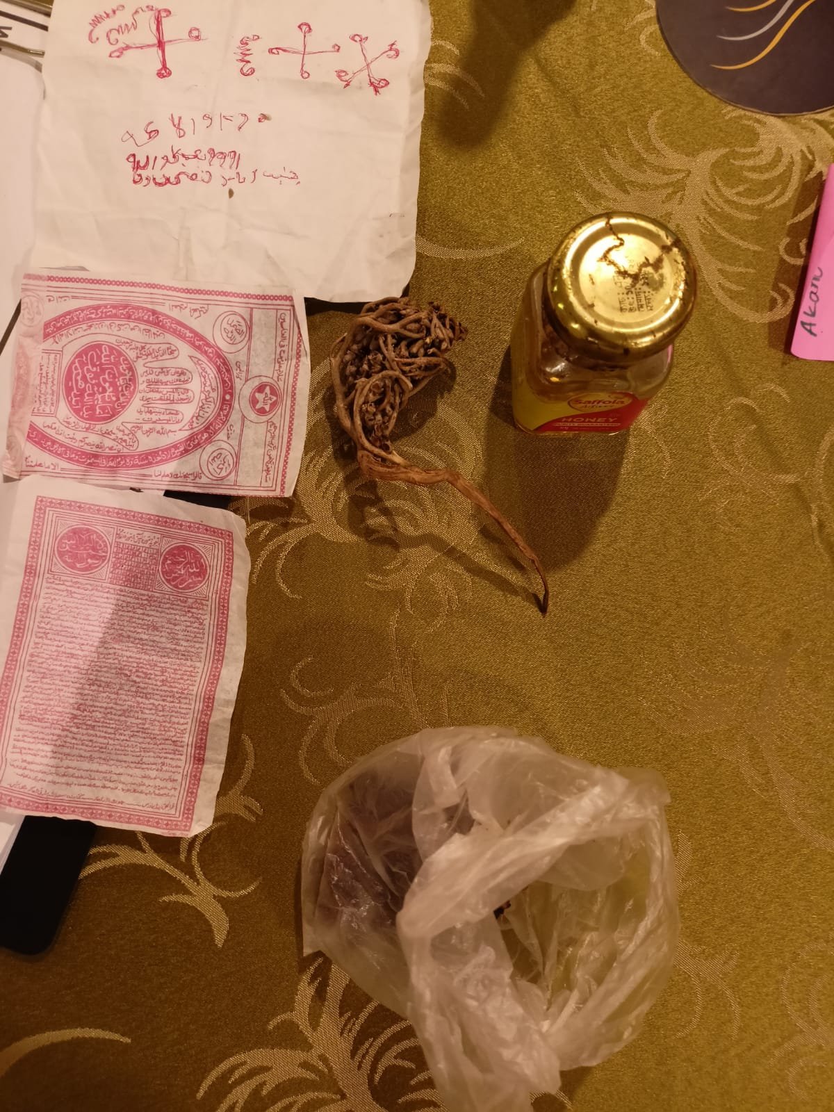 Red printed papers, a white cloth with red markings, a knotted cord, a glass jar and a translucent bag arranged on patterned fabric