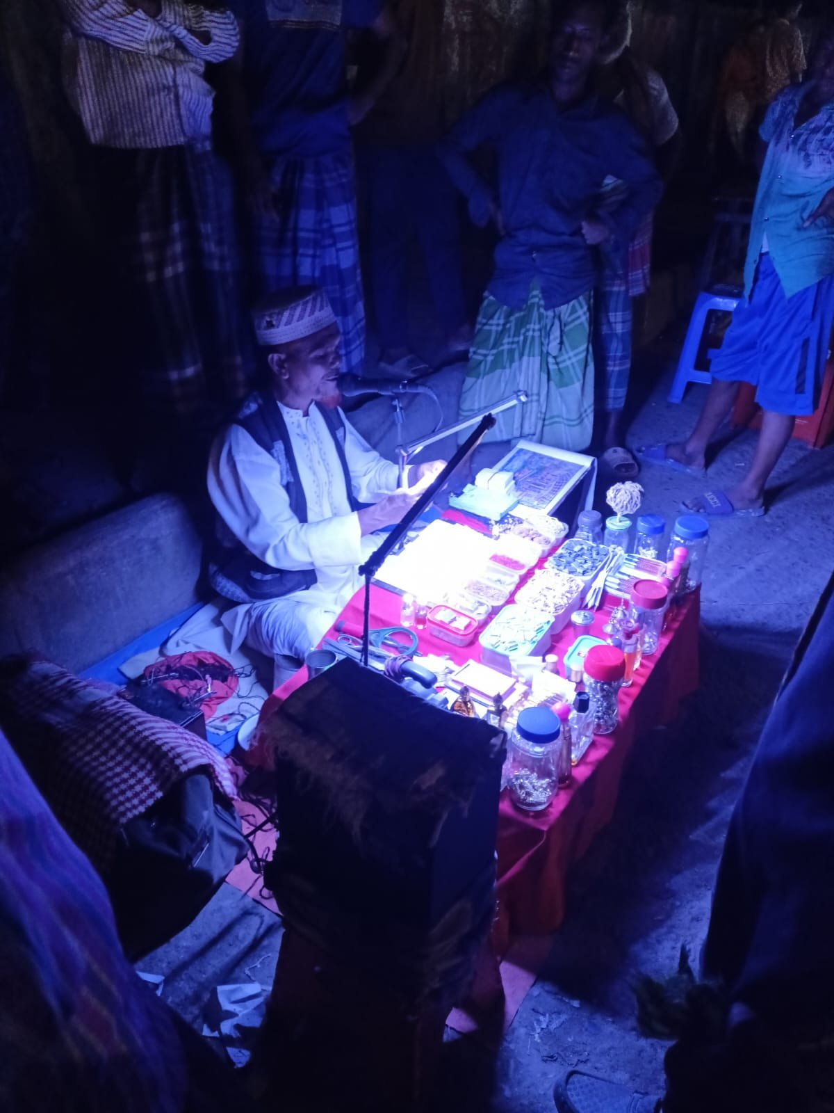 A seated practitioner in white clothing working at a brightly lit table while several adults stand around in a dark setting