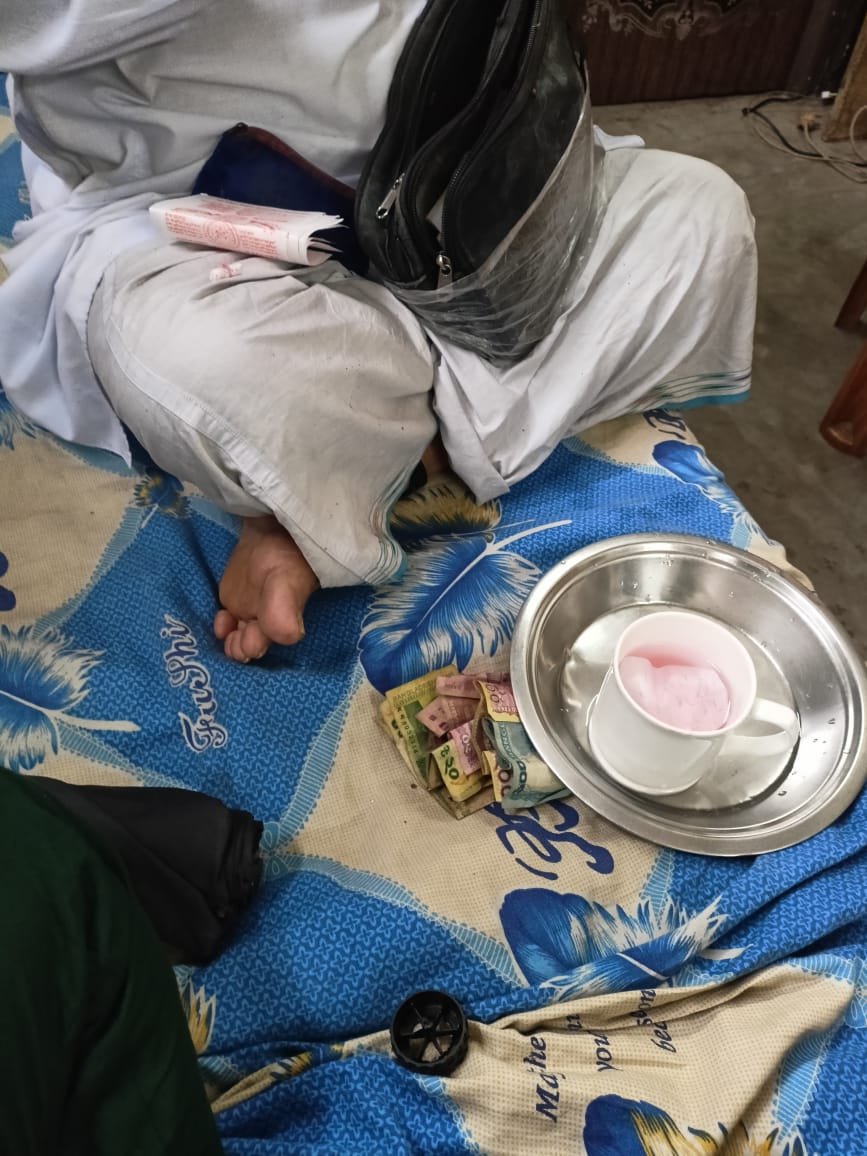 A metal bowl holding a small cup of pink liquid beside folded currency and the lower body of a seated person