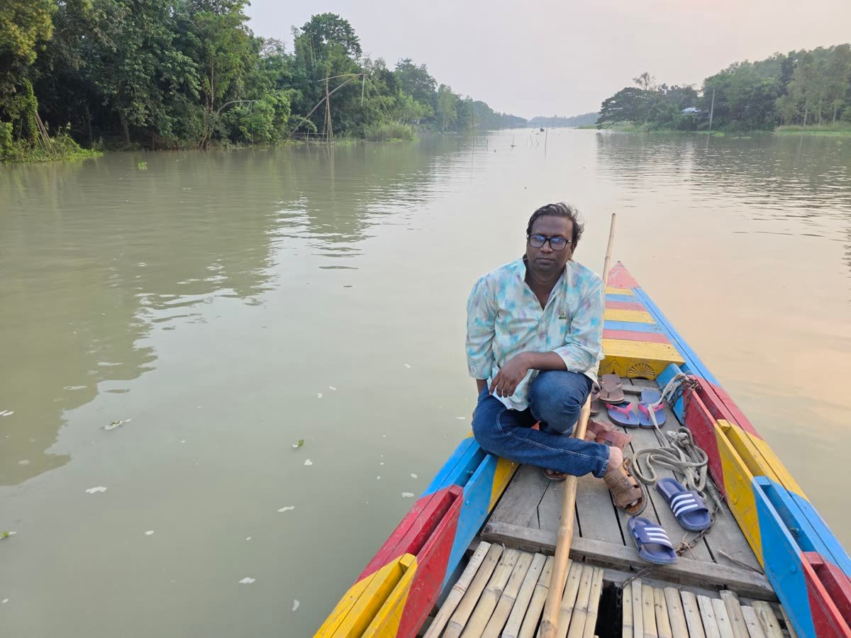 A man sitting quietly at the end of a painted wooden boat on a wide, still river lined with trees